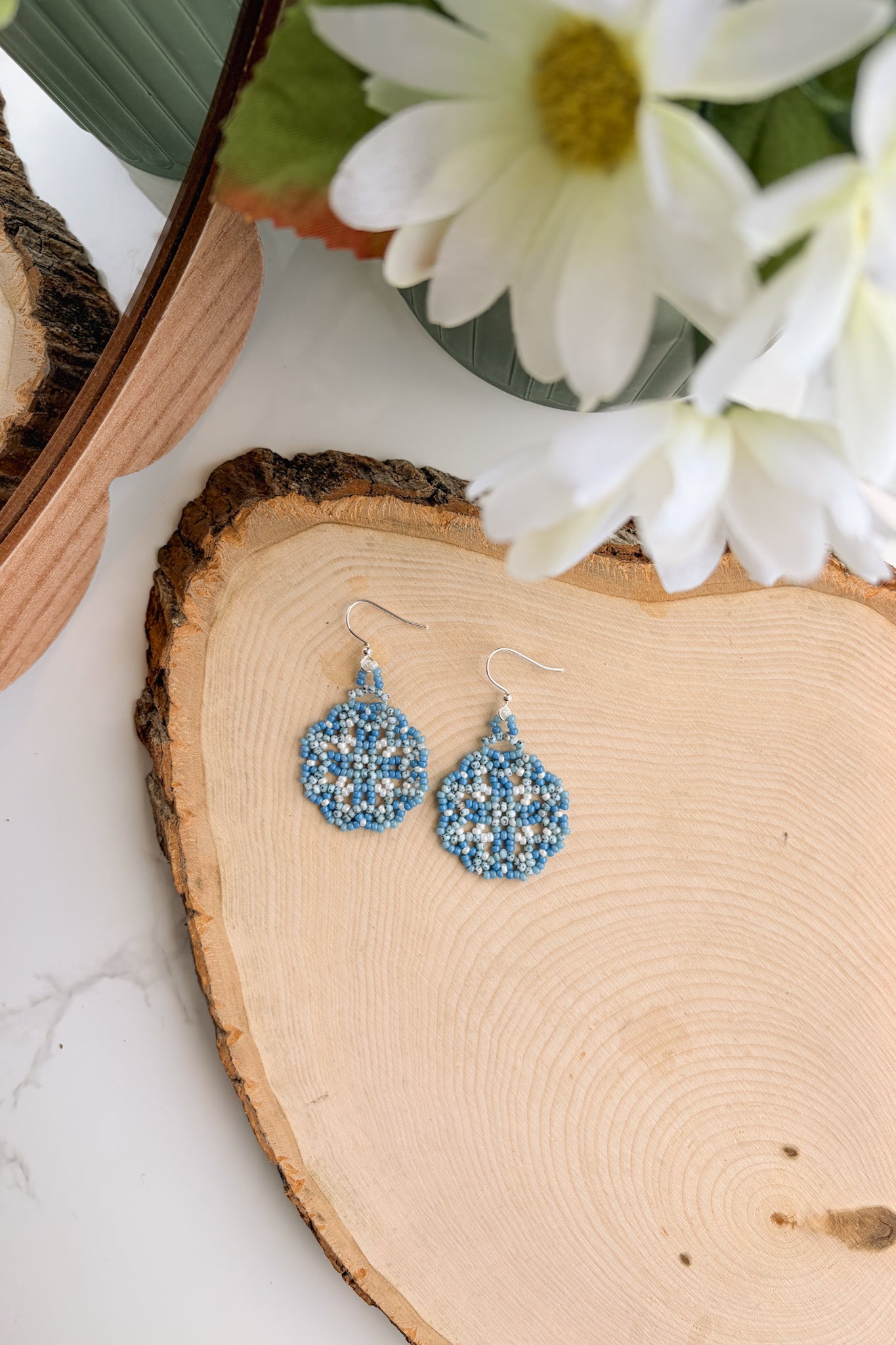 A pair of blue beaded dangle earrings with silver fishhook wires, displayed on a rustic wood slice with white flowers in the background.