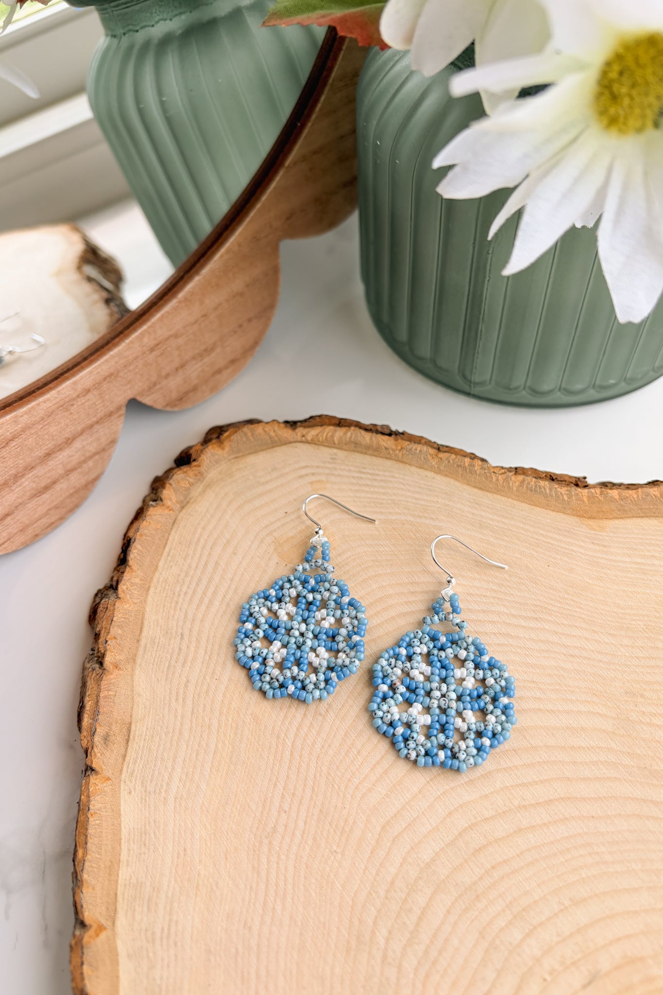Blue and white beaded dangle earrings with silver fishhook wires, displayed on a rustic wood slice with a green vase and white flower in the background.