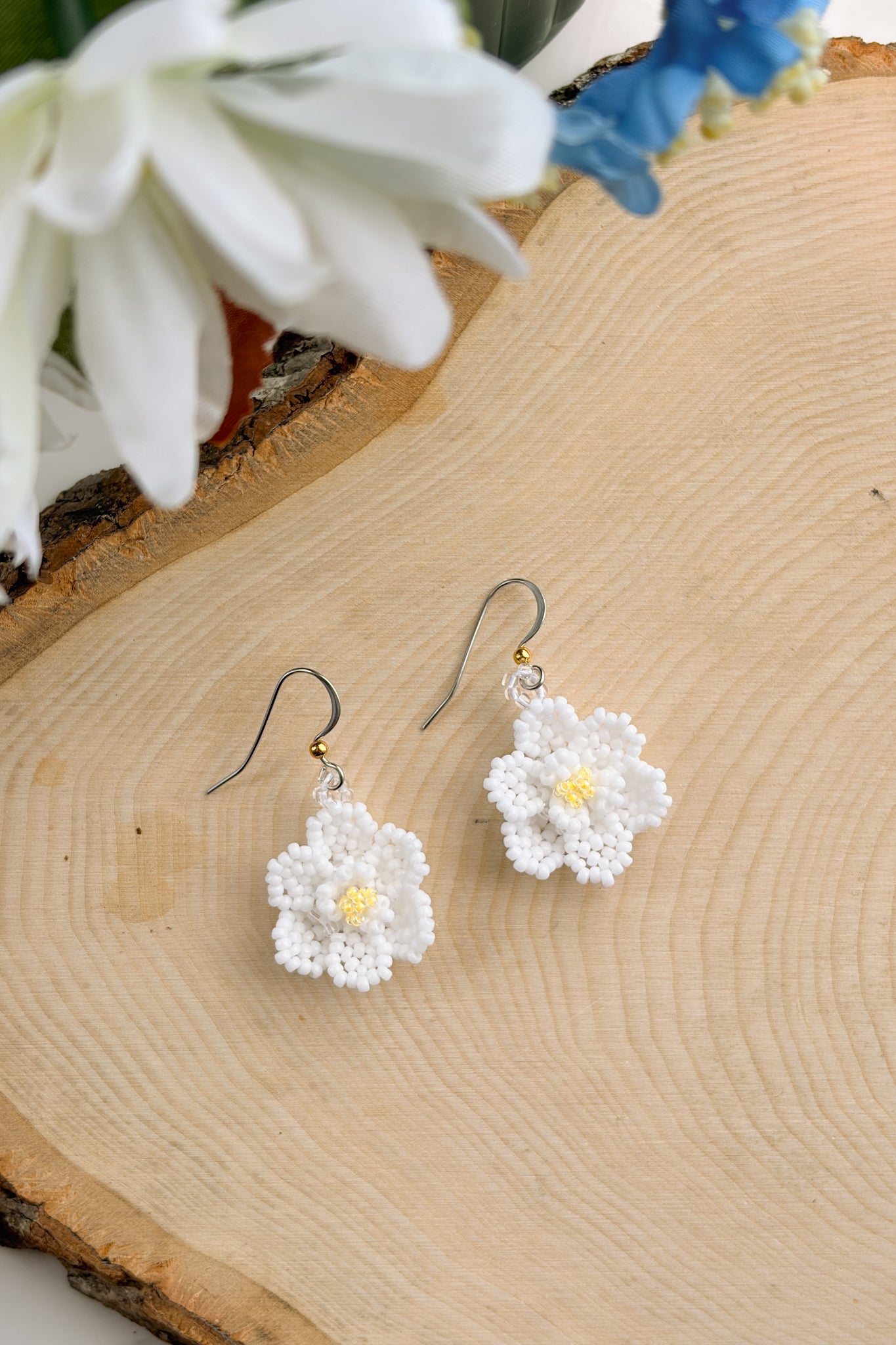 A pair of handmade white flower dangle earrings with yellow centers and silver fishhook wires, displayed on a rustic wood slice background with blurred white and blue flowers.